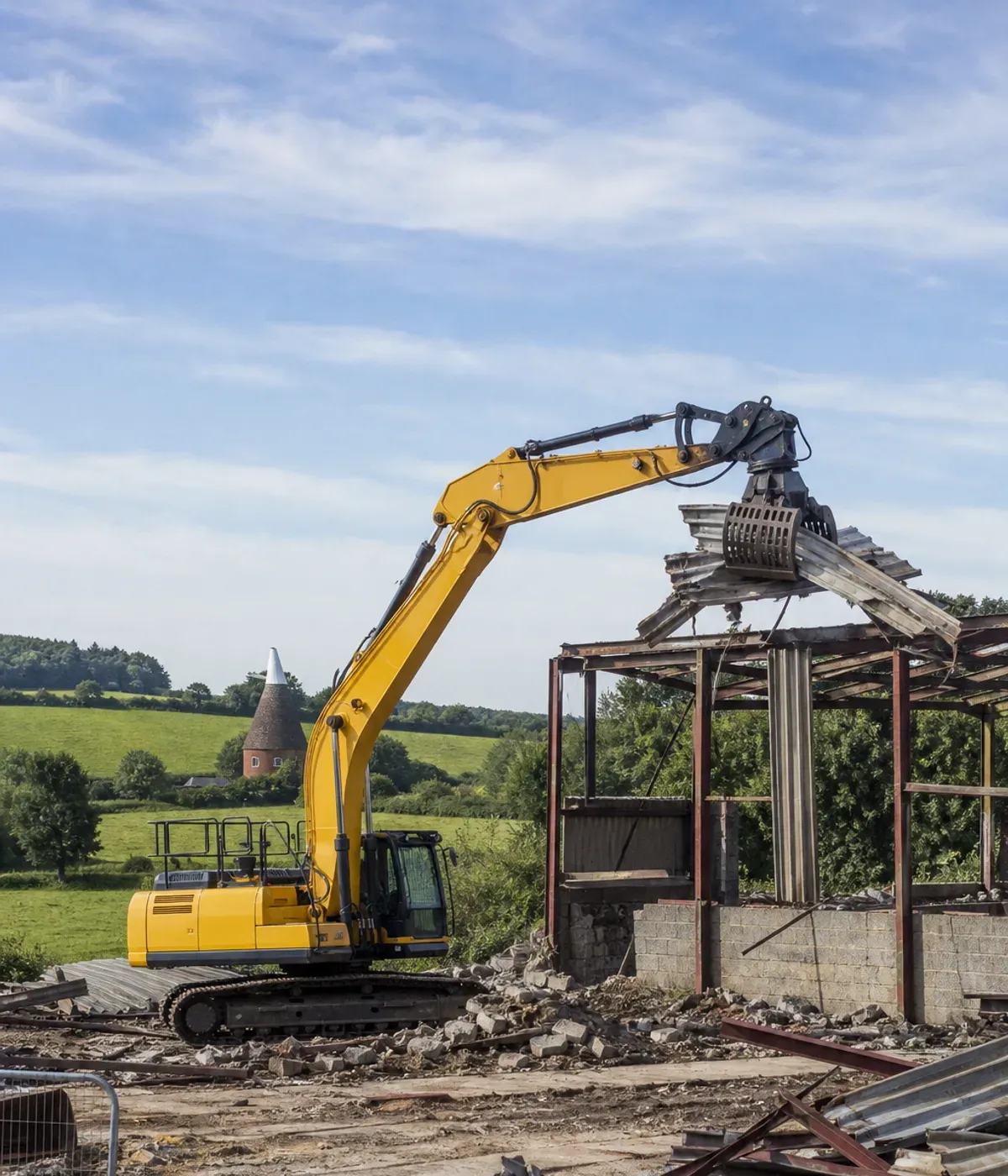 Excavator with a sorting grab dismantling an old agricultural shed, with open countryside and an oast house beyond