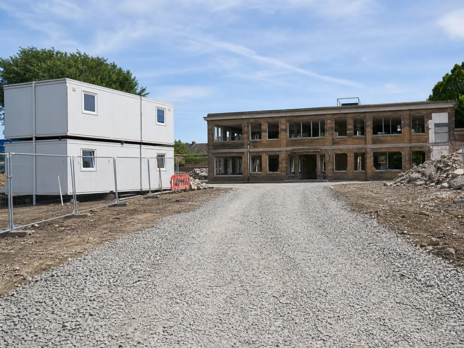 Crushed-stone haul road leading to a vacant brick building, with stacked welfare cabins behind site fencing